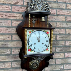 May include: An antique wall clock with a dark wood frame and a white clock face. The clock face features black Roman numerals and red floral accents. The clock has decorative brass elements and a pendulum.