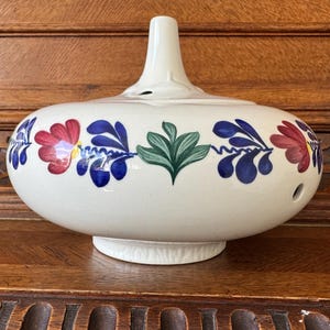 Könnte beinhalten: Eine weiße dekorative Keramikvase mit bauchigem Körper und langem, schlankem Hals. Die Vase ist mit einem sich wiederholenden Blumenmuster in Rot, Blau und Grün verziert. Der Durchmesser des Vasenfußes beträgt ca. 15 cm.
