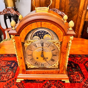 May include: An antique wooden clock with a gold handle and decorative features. The clock face displays Roman numerals and the name "Alexander Cumming". It includes a moon phase indicator. The clock is placed on a red patterned rug.