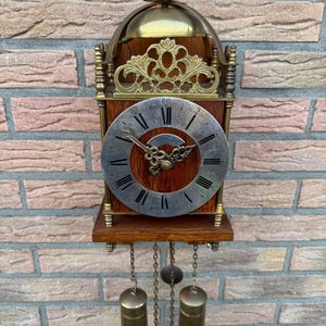 Può includere: Un orologio da parete vintage con cornice in legno e quadrante in metallo. L'orologio presenta una parte superiore decorativa in ottone e pesi a pendolo. Il quadrante dell'orologio ha numeri romani e lancette ornate. L'orologio è montato su un muro di mattoni.