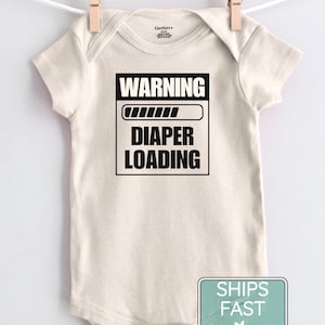 May include: A cream-colored short-sleeve baby bodysuit featuring a black graphic print that reads WARNING with a loading bar icon above the text DIAPER LOADING. The garment has a round neckline and is displayed hanging from a wooden clothespin.