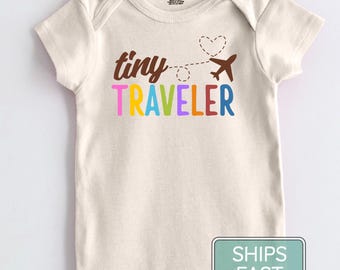 May include: A cream-colored short-sleeve baby bodysuit featuring the brown text tiny traveler with a dashed flight path leading to a heart and a brown airplane. The word traveler is printed in multicolored letters including pink, yellow, blue, green, and red.