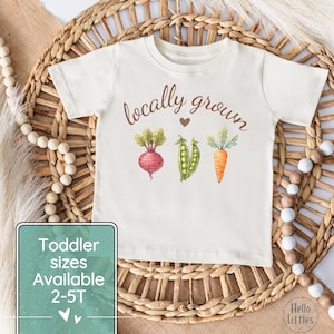May include: Cream-colored toddler t-shirt with the words "locally grown" above a watercolor illustration of a beet, peas, and a carrot. The shirt also has the text "Toddler sizes Available 2-5T".