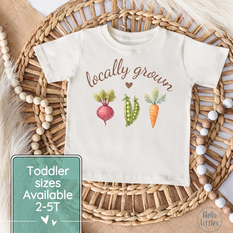 May include: Cream-colored toddler t-shirt with the words "locally grown" above a watercolor illustration of a beet, peas, and a carrot. The shirt also has the text "Toddler sizes Available 2-5T".