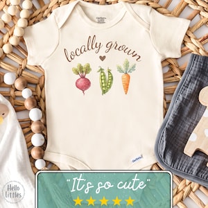 May include: Cream-colored baby onesie with the words "locally grown" in brown script, featuring watercolor illustrations of a beet, peas, and a carrot. The onesie has short sleeves and a round neckline.