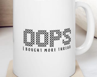 May include: White ceramic mug with the text "oops" in a cross-stitch style font, followed by "I BOUGHT MORE THREAD". The mug has a curved handle and sits on a square coaster. The background includes wooden elements and a bowl with fruit.