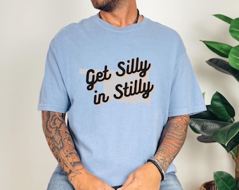Könnte beinhalten: Hellblaues T-Shirt mit dem Text "Get Silly in Stilly" in schwarzer Schrift. Das T-Shirt zeigt eine graue Umrisslinie des Bundesstaates Oklahoma. Das T-Shirt ist aus einem weichen Material gefertigt.