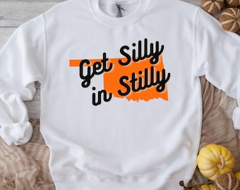 Könnte beinhalten: Weißes Sweatshirt mit dem Text "Get Silly in Stilly" in schwarzer Schrift und einer orangefarbenen Umrisszeichnung von Oklahoma. Ein lässiges Kleidungsstück.