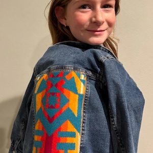 May include: A blue denim jacket with a colorful geometric pattern on the back. The pattern is in shades of red, orange, yellow, teal, and white.