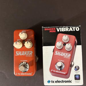 Puede incluir: Un pedal de efectos de guitarra Shaker Mini Vibrato naranja con perillas blancas y el texto "SHAKER MINI VIBRATO". El pedal está junto a su caja, que también muestra el pedal y el texto "SHAKER VIBRATO MINI".