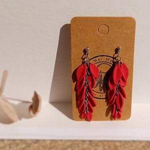 Op de afbeelding: Rode hangende oorbellen met een ketting en meerdere blaadjesvormige bedels. De oorbellen worden getoond op een bruine kaart met de tekst "Mar Mo" en een logo.