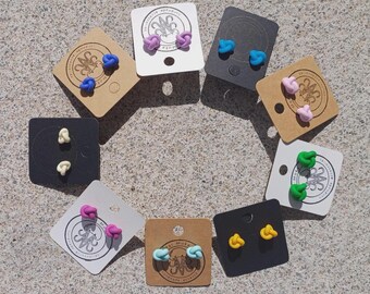 May include: A collection of eight pairs of knot-shaped stud earrings displayed on individual square cards. The earrings come in various solid colors including purple, blue, pink, cream, green, magenta, light blue, and yellow, each on branded cards.
