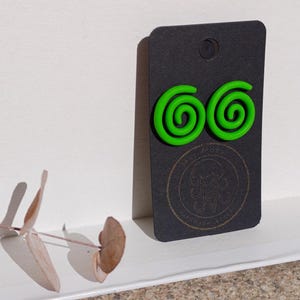 Op de afbeelding: Een paar groene spiraalvormige oorbellen op een zwarte kaart met de tekst "Mind Over Matter" in een cirkel.