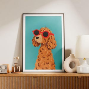 Puede incluir: Una lámina de arte mural enmarcada que muestra a un caniche marrón de pelo rizado con gafas de sol redondas de color rojo brillante sobre un fondo de color verde azulado sólido. La ilustración se presenta en un marco negro fino.