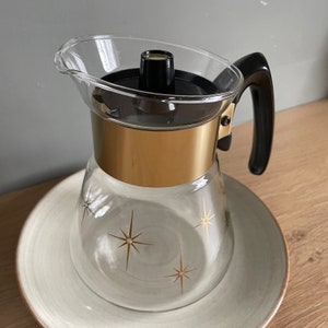 May include: A glass coffee pot with a gold band and a black handle. The pot has a clear glass body with a starburst design and sits on a matching saucer.