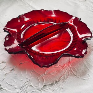 May include: A divided serving dish made of ruby red glass with a scalloped edge. The dish has a raised centre divider.