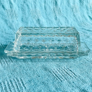 May include: A clear glass butter dish with a diamond pattern. The dish is rectangular with a lid and small handles. The butter dish is made of glass and sits on a blue surface. The dimensions are approximately 15 cm in length.