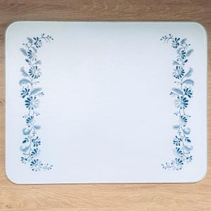May include: White rectangular chopping board with rounded corners. It has a floral design in shades of blue and grey, arranged vertically along the left and right edges. The background is a solid white colour.