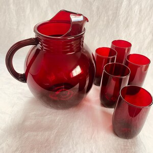 Peut inclure: Un pichet en verre rouge avec un corps arrondi et une poignée, accompagné de six verres rouges assortis. Le pichet a un bec verseur. L'ensemble de verrerie est probablement destiné à servir des boissons.