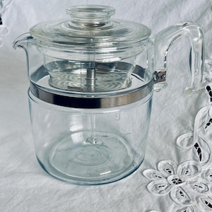 May include: A clear glass coffee percolator with a glass lid and handle. The percolator has a silver-coloured metal band around the middle and a clear glass interior. The handle is curved and attached to the side. The percolator is on a white surface.