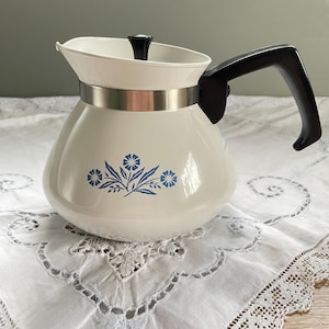 May include: Cream-coloured coffee pot with a black handle and lid. The pot has a silver band and a blue floral design. It sits on a white, embroidered tablecloth. A vintage kitchenware item.