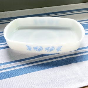 Peut inclure: Un plat à four rectangulaire blanc cassé aux coins arrondis, orné d'un motif floral bleu sur le bord. Le plat vide est posé sur une surface à rayures bleues et blanches.