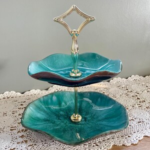 May include: Two-tiered serving tray with a teal and brown glazed finish. The scalloped edges of the ceramic plates are accented with a brown trim. A gold-toned metal handle and central support connect the tiers, creating a decorative display piece.