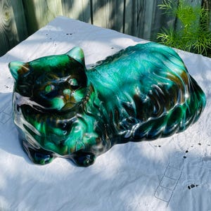 May include: A ceramic cat ornament in shades of green and brown. The glossy finish highlights the details of the cat's fur and features. The cat is lying down, with its head and tail slightly raised. A decorative item.