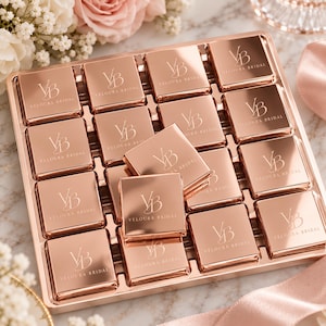 May include: A square rose gold tray containing sixteen individual square rose gold pieces. Each piece is engraved with the letters VB and the text VELOURA BRIDAL. The pieces are arranged in a grid, with one piece placed diagonally on top.
