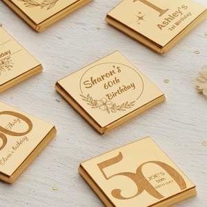 May include: Gold-wrapped chocolate bars with engraved birthday messages. The bars display text including "Sharon's 60th Birthday," "Ashley's 1st Birthday," and "Joe's 50th Birthday." Floral and number designs are also featured on the bars.