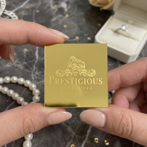 以下が含まれることがあります： 「Prestigious Jewellers」の文字と虎のエンブレムが入った金の正方形が両手に持たれています。真珠のネックレスと白い箱に入った指輪が背景にあります。