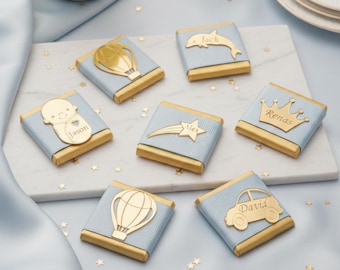 May include: Six square chocolate bars wrapped in light blue ribbed paper with gold foil edges. Each bar features a gold metallic cutout: a hot air balloon, a dolphin with the name Jack, a baby with the name Jason, a shooting star with the name Alex, a crown with the name Renas, and a car with the name David.