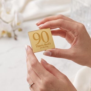 以下が含まれることがあります： 「90」の数字と「Minnie Hunt's 90th Birthday」の文字が刻まれた金色の正方形が、人の手で持たれています。これは、記念品であり、おそらく特別な誕生日のためのものです。