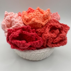 May include: A crocheted flower arrangement in a white crocheted bowl. The flowers are in shades of pink, orange, and red.