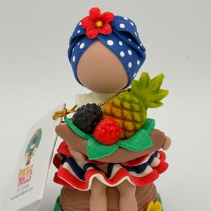 May include: A decorative doll figurine featuring a blue head wrap with white polka dots and a red flower. The doll holds a tray of fruit, including a pineapple, blackberries, and raspberries. The doll's skirt has red, white, and blue ruffles. The base is brown and green.