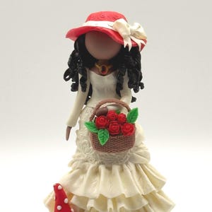 Può includere: Statuina artigianale di una donna con abito color crema e cappello rosso con fiocco bianco. Tiene in mano un cestino di rose rosse. Un fungo rosso con pois bianchi e fiori ai suoi piedi.