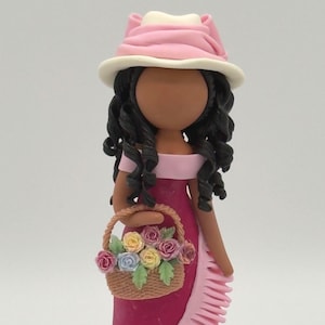 May include: A handcrafted figurine of a woman in a pink dress and hat, holding a basket of flowers. The dress has a ruffled design, and the hat is white with a pink brim. The figurine has dark curly hair and a brown complexion.