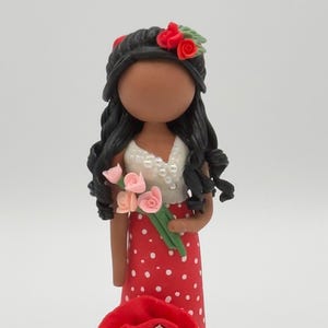 May include: A handcrafted figurine of a woman in a red polka-dot dress, holding pink flowers. She has black hair adorned with red roses and a matching red flower at her feet. The figurine is approximately 6 inches tall.