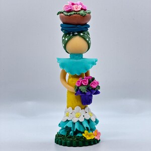 May include: A handmade doll figurine with a yellow dress, blue and white floral accents, and a green base. The doll is holding a bouquet of pink flowers and has a green and blue head wrap with pink flowers on top.