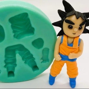Op de afbeelding: Groene siliconen mal met verschillende vormen om een taarttopper te maken, waaronder een figuur met volledig lichaam, benen, armen en een hoofd. De mal bevindt zich naast een afgewerkte taarttopper van een cartoonfiguur met stekelig zwart haar, een oranje jumpsuit en blauwe laarzen.