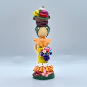 May include: A handmade doll figurine with a green and white patterned head wrap, a yellow dress with orange ruffles, and a bouquet of colorful flowers.