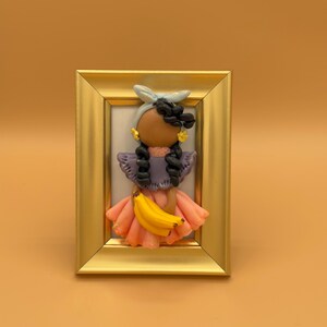 May include: A framed artwork featuring a clay figure of a person with dark braids, wearing a lavender top, peach skirt, and holding a bunch of bananas. The figure has a light blue headscarf and gold earrings. The frame is gold.