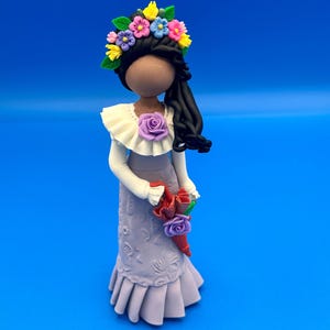 May include: A handcrafted figurine of a woman with dark hair, wearing a lavender dress and a white blouse with a ruffled collar. The figurine has a floral crown and holds a bouquet of red and purple flowers. The background is a solid blue.
