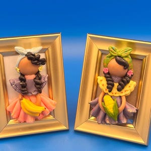 May include: Two framed clay figurines of women in traditional attire. One holds a banana, the other a corn cob. Both have dark hair, braids, and wear colorful dresses. The frames are gold, set against a blue background.