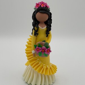 Può includere: Statuina artigianale di una donna in abito giallo con gonna a balze, con in mano un cestino di fiori rosa. La statuina ha capelli neri, una corona di fiori e una collana di perle. La base è bianca.