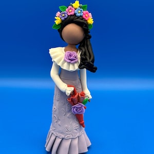 Può includere: Statuina artigianale di una donna con una corona floreale e un lungo vestito color lavanda. La figura tiene in mano un bouquet di fiori rossi e viola. La bambola ha i capelli neri e una camicetta bianca con colletto a balze.