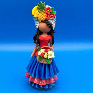 May include: A handcrafted figurine of a woman in a red top and blue skirt, carrying a basket of flowers. Her head is adorned with a turban decorated with a pineapple, bananas, and red flowers. The figurine is set against a solid blue background.
