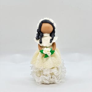 May include: A white porcelain doll wearing a white dress with lace trim and a white flower crown. The doll has black hair and brown eyes and is holding a bouquet of white flowers.