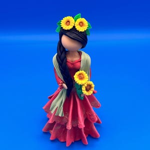 May include: A handcrafted figurine of a woman in a red dress, adorned with a sunflower crown and holding sunflowers. The figure has a long black braid and a green shawl. The background is a solid blue.