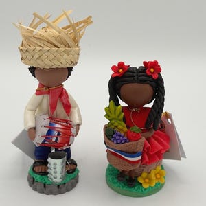 May include: Two handcrafted figurines. One figure wears a straw hat, white shirt, red scarf, and blue pants, holding a drum. The other figure wears a red dress and holds a basket of fruit. Both figures are on a green base.